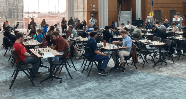 Several rows of tables in the Atrium at Central Library with multiple people of all ages seated playing chess.