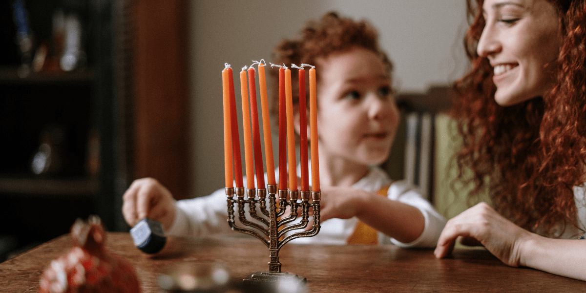 A mother and son sit at a table with a Menorah. The boy is holding a small dreidel.