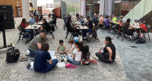 Several rows of tables in the Atrium at Central Library with multiple people of all ages seated playing chess.